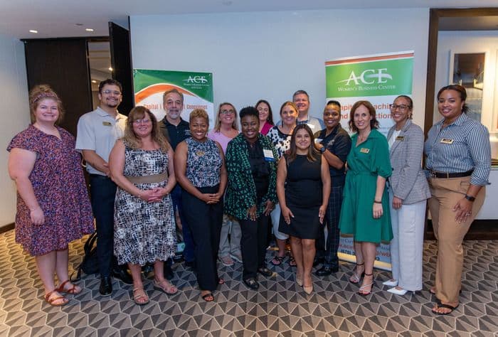 A diverse group of professionals stands together indoors, smiling next to two ACE branded banners.