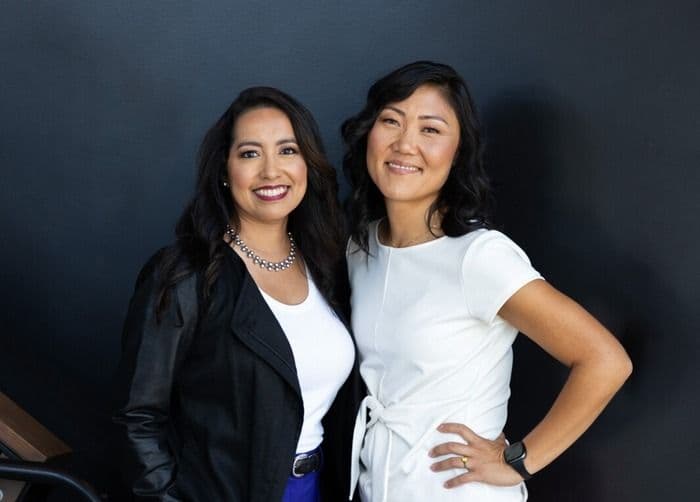 Two diverse professional women, both smiling, stand confidently against a dark background.