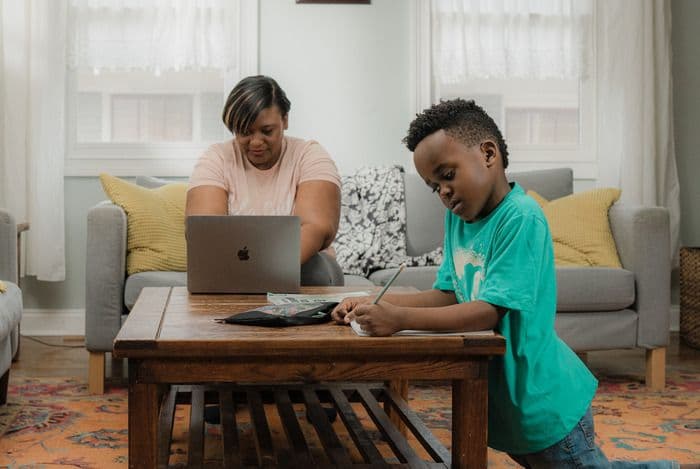 Woman works on laptop while young boy writes on coffee table in living room.