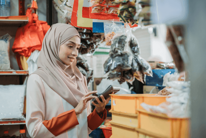 Young woman in hijab using smartphone amidst goods in a bustling market.