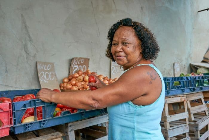 Smiling woman at market stall arranging fresh tomatoes and onions for sale.
