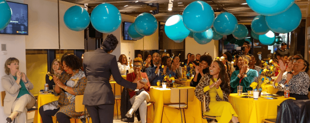A speaker addresses a large audience, mostly women, clapping under teal balloons.