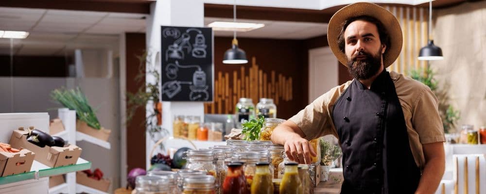 Bearded man in a hat and apron leans on a counter in a specialty food store.