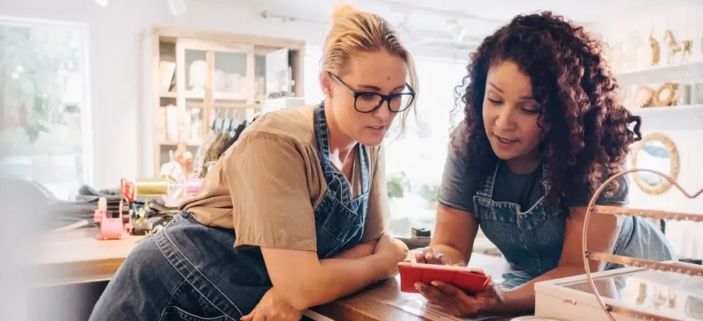Two women in aprons look intently at a red smartphone in a bright small shop.