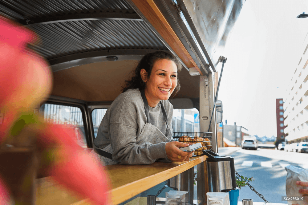 Smiling woman in apron serves a customer from her food truck window on a sunny day.
