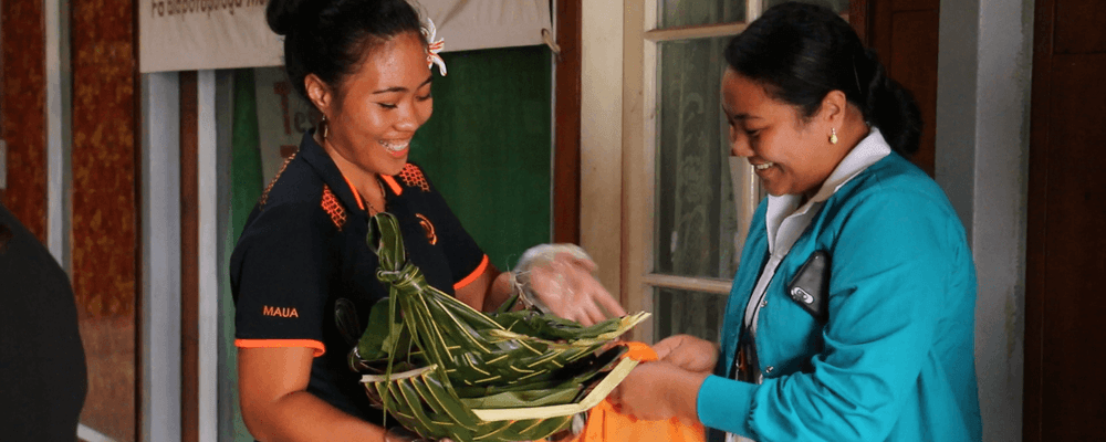 Two smiling women interact, one holding a woven basket of green leaves, possibly traditional goods.