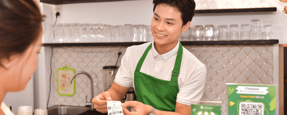 Smiling barista in green apron hands a receipt to a female customer at a cafe counter.