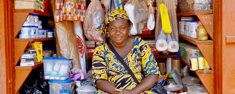 A smiling woman in a colorful dress sits inside her small, well-stocked shop.