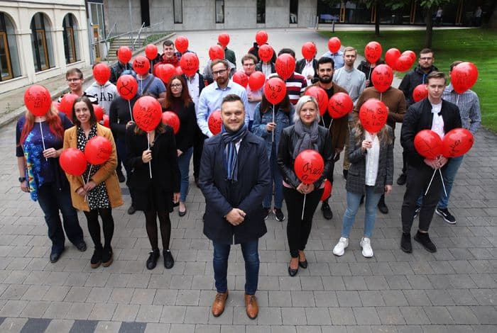 A large group of people standing outdoors, many holding bright red balloons.