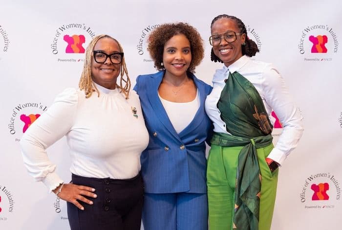 Three smiling women of color, in professional attire, pose in front of an event backdrop.
