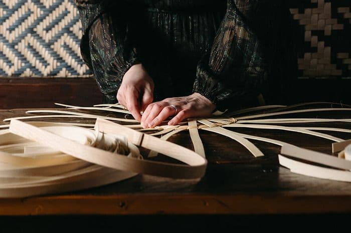 Close-up of hands meticulously weaving light-colored strips into an intricate pattern.
