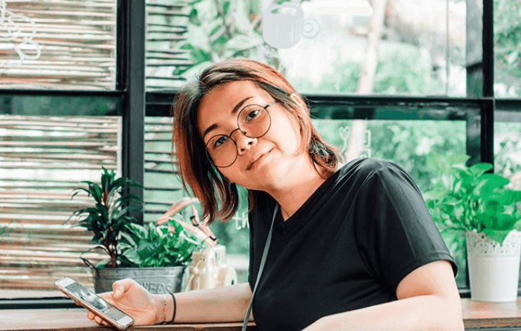 A woman wearing glasses and a black shirt holds a phone in a cafe.