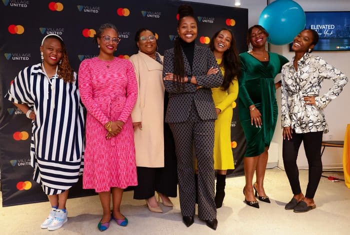 Seven women in stylish outfits pose confidently before a black Mastercard branded backdrop.