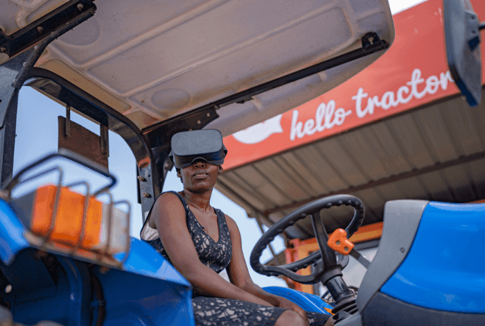Woman in a VR headset sits in a blue tractor, with a 'hello tractor' sign in background.
