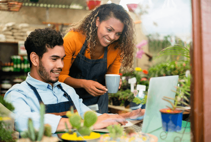 Two smiling florists review laptop content in a vibrant plant nursery.