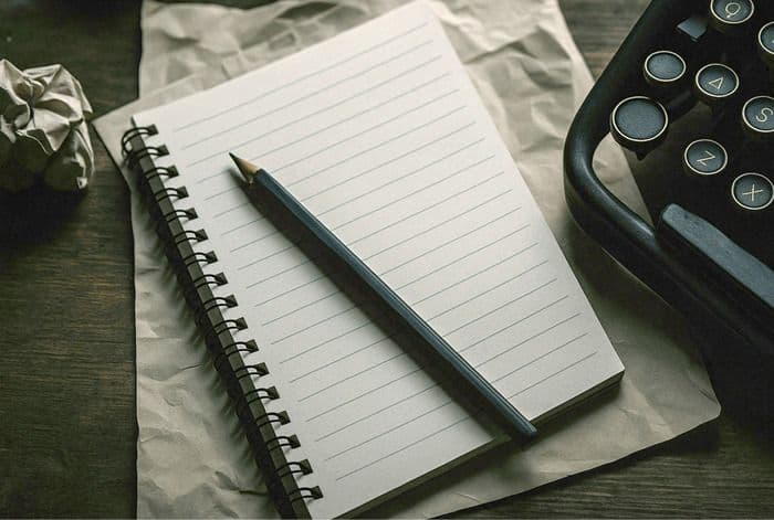 Spiral notebook, pencil, crumpled paper, and vintage typewriter on a dark wooden surface.