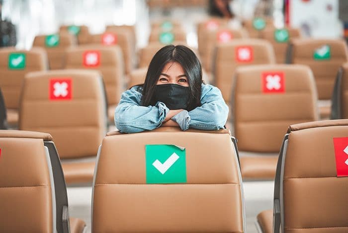 Young woman in mask smiles behind airport chair with social distancing markers.