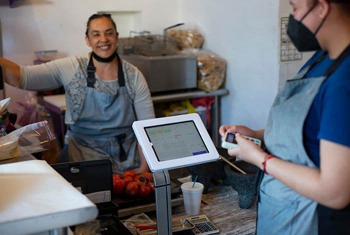 A smiling vendor stands behind a counter while a customer uses a payment terminal.