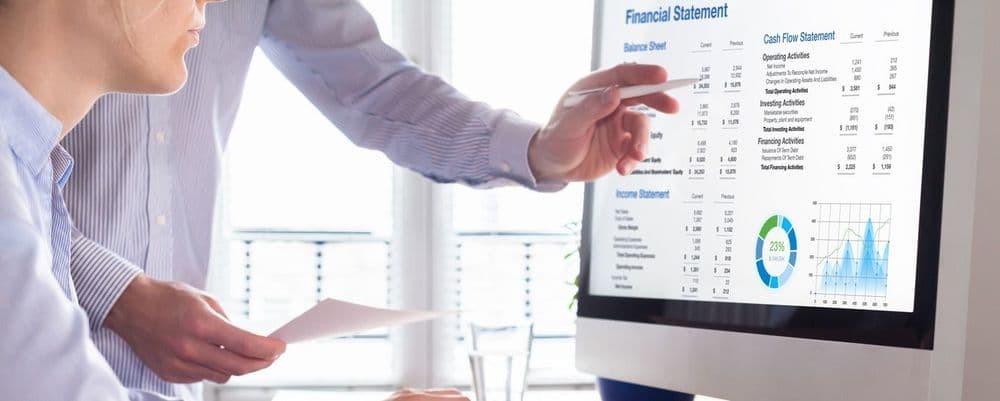 Two individuals review a financial statement displayed on a monitor, one pointing at data.