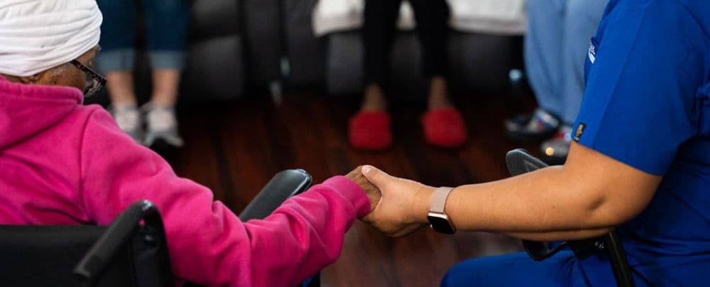 A patient in a pink hoodie in a wheelchair holds hands with a caregiver.