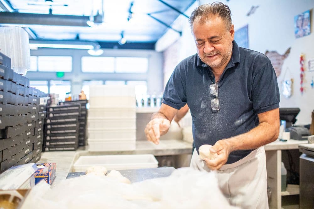 A man in a blue shirt and white apron kneads dough in a busy bakery.