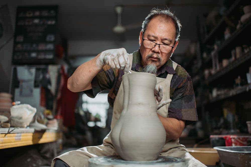 An older man with glasses sculpts a tall clay vase on a pottery wheel.