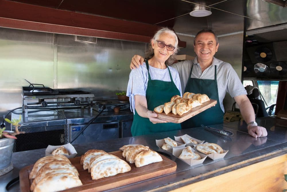 Senior couple in aprons proudly display fresh empanadas from their food truck.