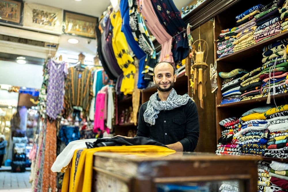 A man in a keffiyeh smiles behind a counter in a vibrant, textile-filled clothing shop.