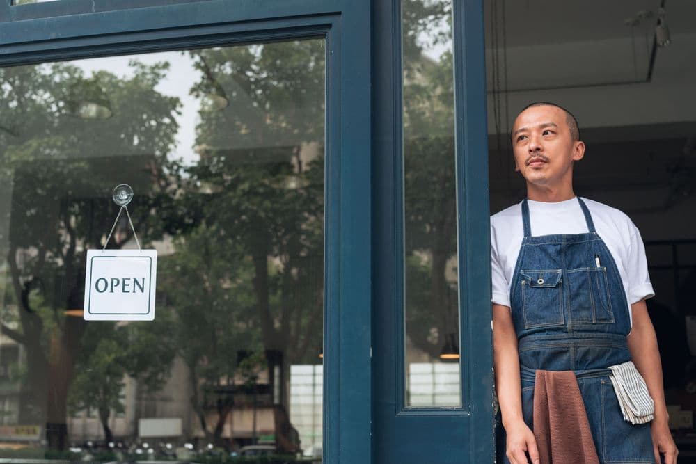Man in denim apron stands in open doorway next to an 'OPEN' sign.