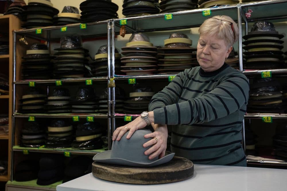 Woman shaping a grey hat in a workshop filled with shelves of various hats.