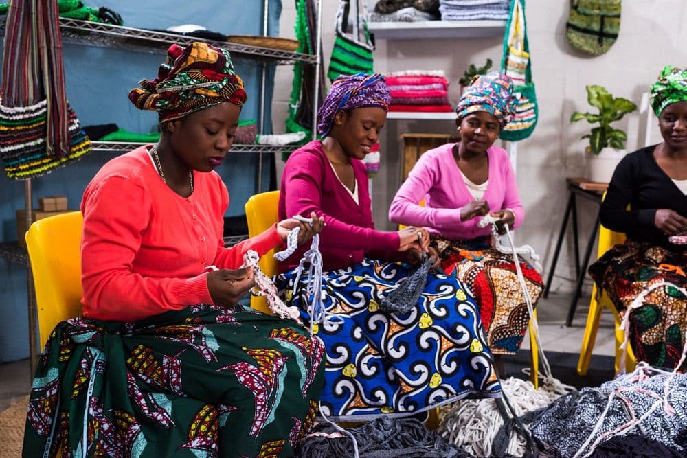 Four women in colorful head wraps and skirts craft white items in a workshop setting.