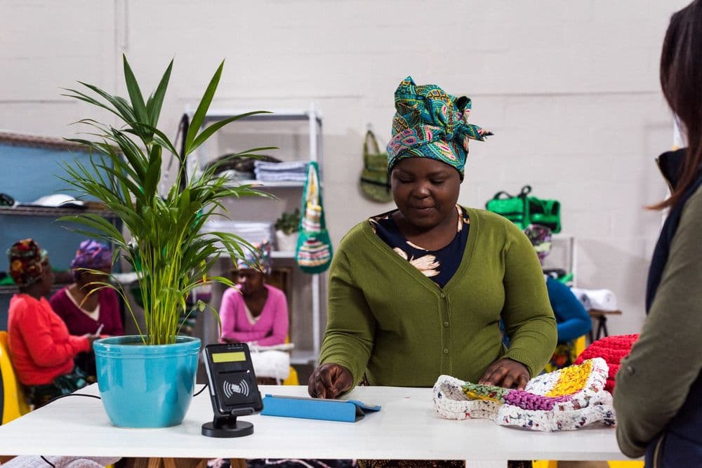 Woman in green sweater and colorful headwrap examines handmade crafts at a shop counter.
