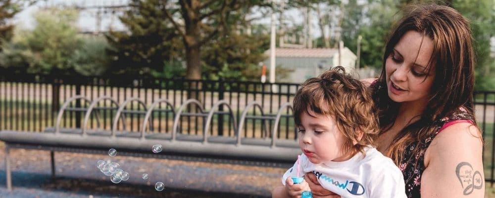 A woman holds a young child outdoors who is watching bubbles float by.