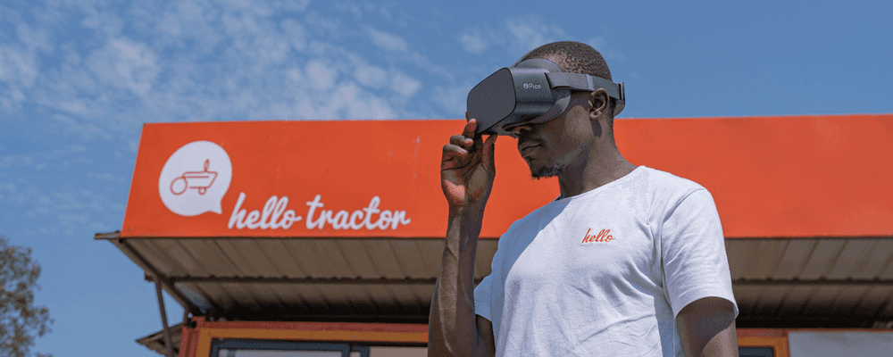 A man wearing a VR headset adjusts it, standing near a 'hello tractor' sign.