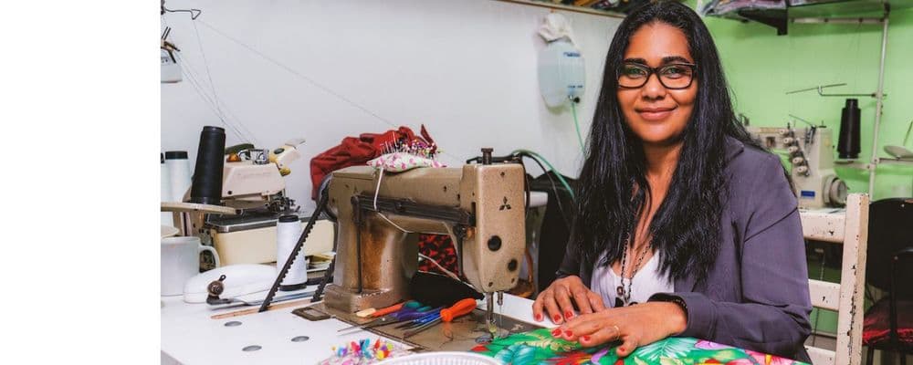 A smiling woman with glasses works at a sewing machine, creating colorful fabric items.