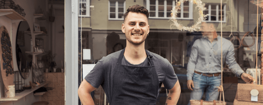 A smiling man wearing an apron stands proudly in his small business storefront.