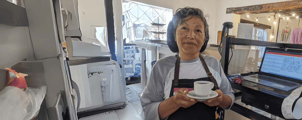 A smiling woman in an apron holds a small cup of coffee in an indoor setting.