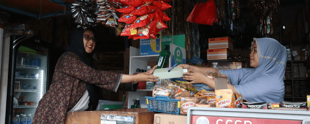 Two smiling women in hijabs exchange an item over a counter in a busy small shop.