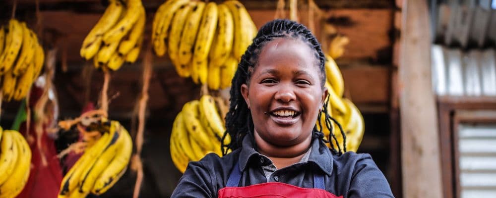 Smiling woman in black shirt and red apron stands before hanging bunches of ripe bananas.