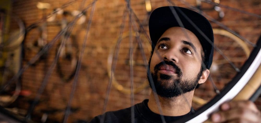 Man with beard and cap looks up, holding a bike wheel in a repair shop.