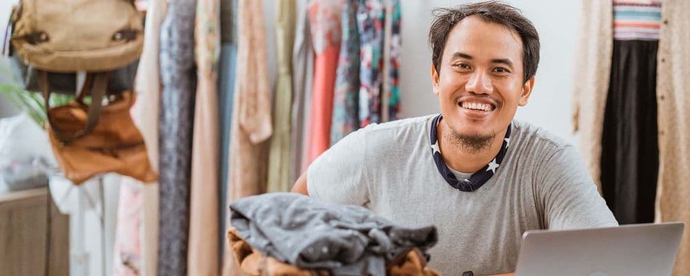 Happy man in grey shirt smiles, working on a laptop in a clothing shop.
