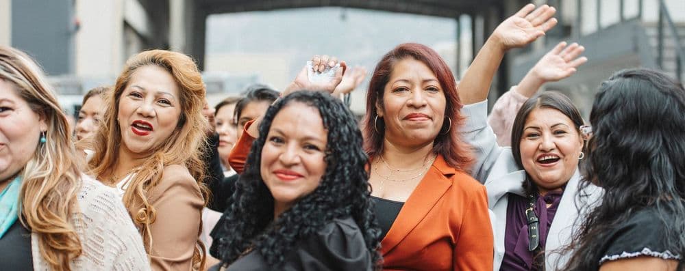 Group of diverse women smiling and waving energetically in an outdoor setting.