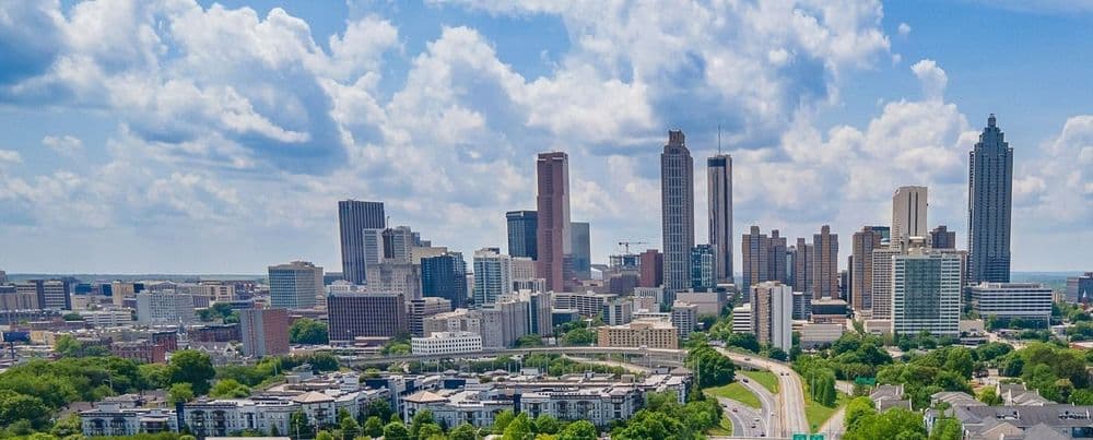 Sprawling city skyline featuring numerous tall buildings, green trees, and cloudy blue sky.