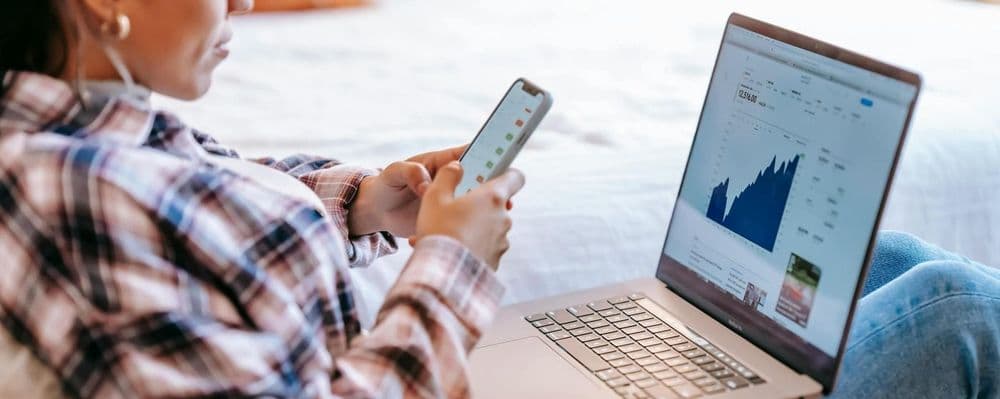 Person in plaid shirt reviewing data on phone and laptop showing charts.