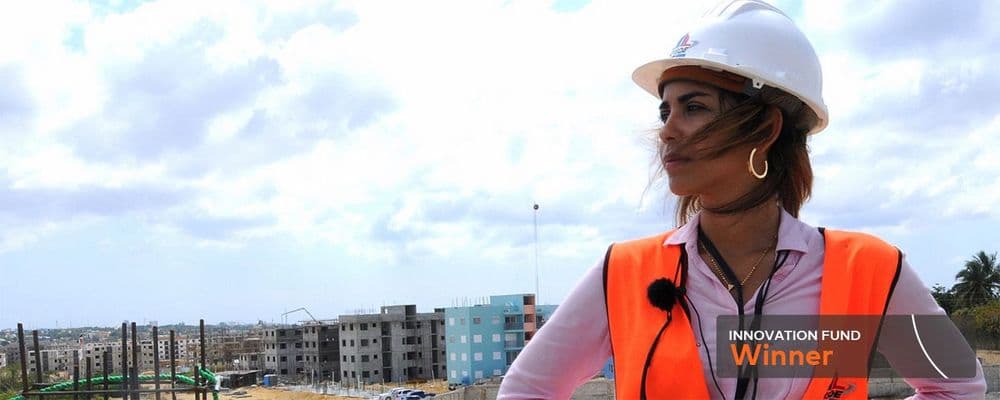 Woman in white hard hat and orange vest, labeled 'Winner', overlooks a construction site.