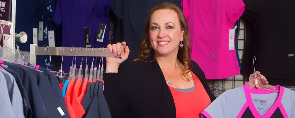 A smiling woman stands in a retail store, holding a clothes rack displaying medical scrubs.