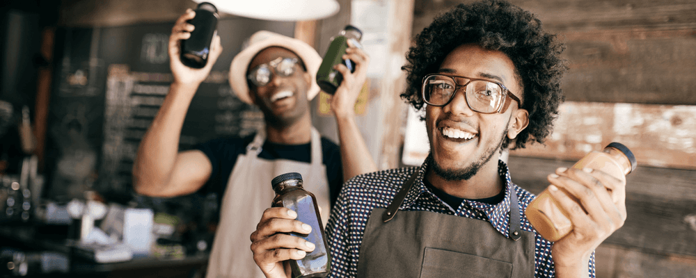 Two smiling men in aprons enthusiastically hold up bottled beverages in a cafe.