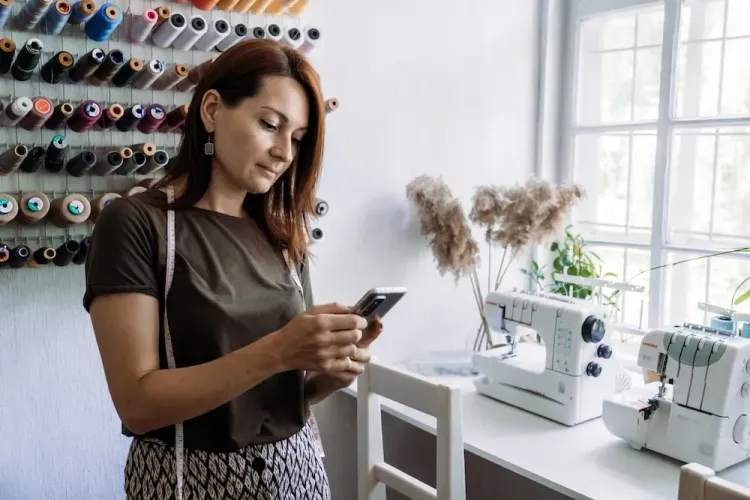 Woman checks phone in a bright sewing room with spools of thread and machines.
