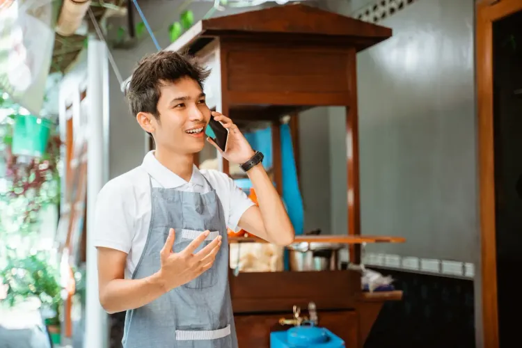 A smiling young man in an apron talks on his phone near a food cart.