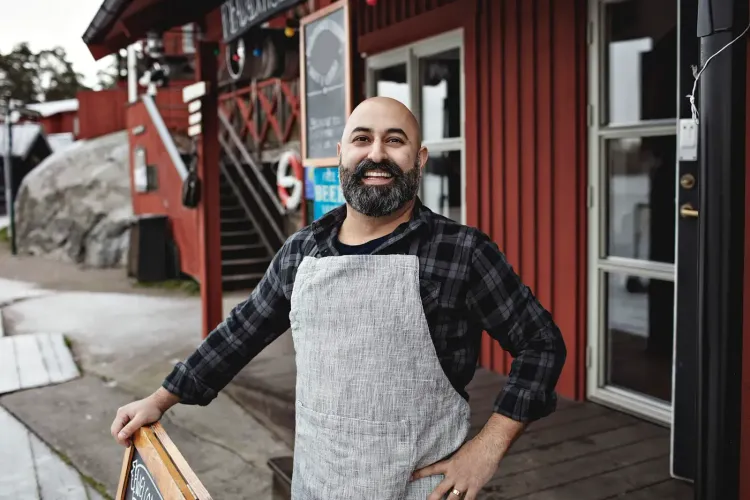 A smiling man with a beard and apron stands outside a red building.